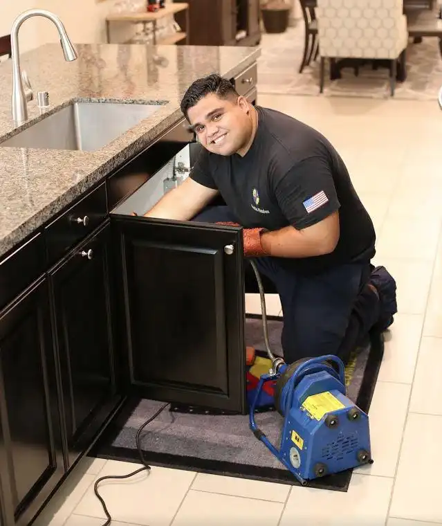 A Guardian Plumbers, Heating, Air & Electrical technician, with a friendly smile, is cleaning out a kitchen drain line using a professional drain machine. His positive demeanor and the efficient equipment demonstrate the thorough and customer-friendly service provided by Guardian Plumbers, Heating, Air & Electrical.
