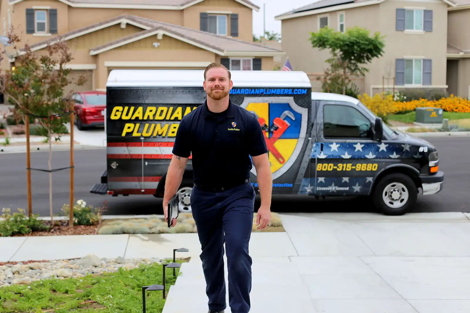 A Guardian Plumbers, Heating, Air & Electrical technician, equipped with tools, walks up to a customer's home, exuding professionalism and readiness. The plumbing van, prominently displaying the Guardian Plumbers, Heating, Air & Electrical logo, is parked behind him, signaling reliable and prompt service is about to be delivered.