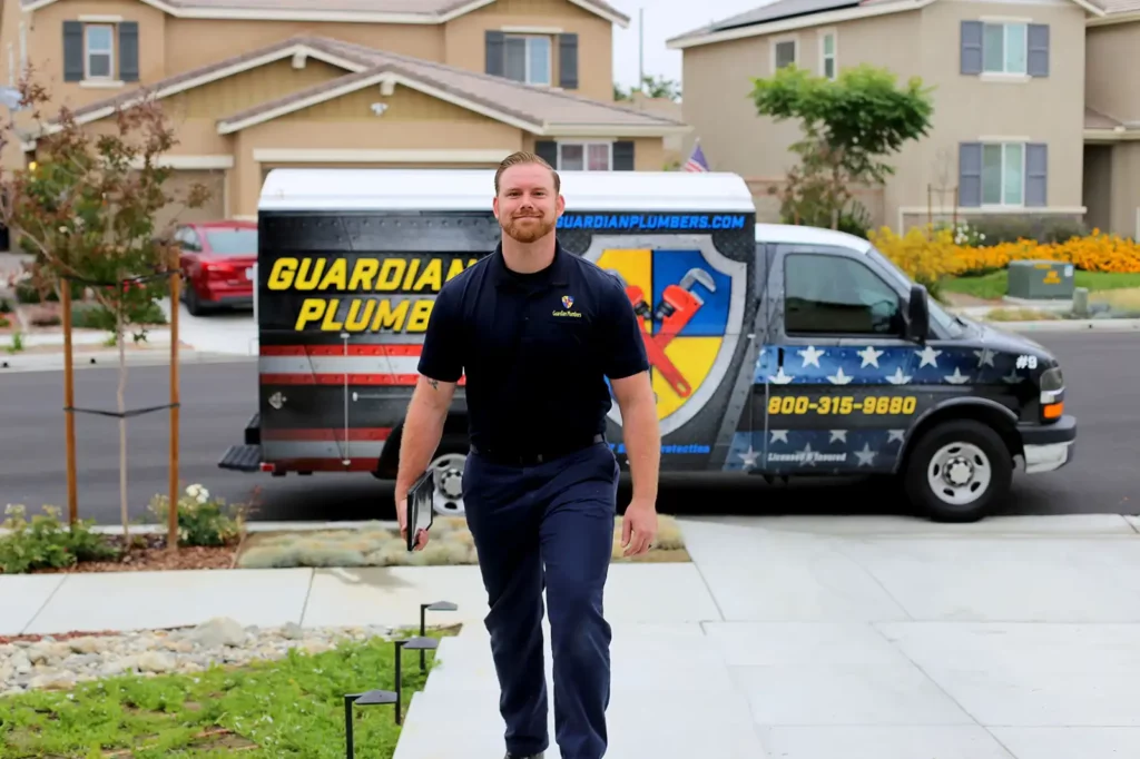A Guardian Plumbers, Heating, Air & Electrical technician, equipped with tools, walks up to a customer's home, exuding professionalism and readiness. The plumbing van, prominently displaying the Guardian Plumbers, Heating, Air & Electrical logo, is parked behind him, signaling reliable and prompt service is about to be delivered.